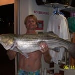 Man proudly holds a large striped bass fish indoors.