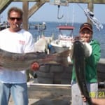 Two people proudly holding large fish on a dock by the water.